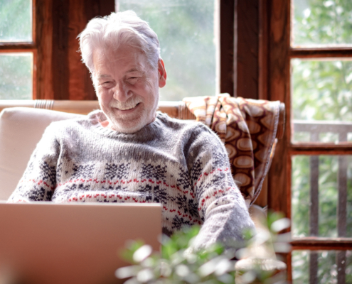 Front view, smiling senior man in winter sweater sitting in living room using laptop computer