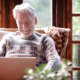 Front view, smiling senior man in winter sweater sitting in living room using laptop computer